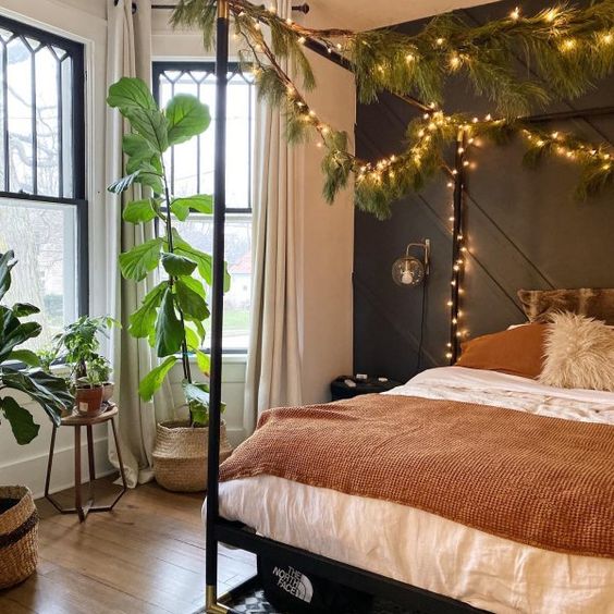 a cozy bedroom with a bow window, a black accent wall, a canopy bed decorated with fur branches and lights plus statement potted plants