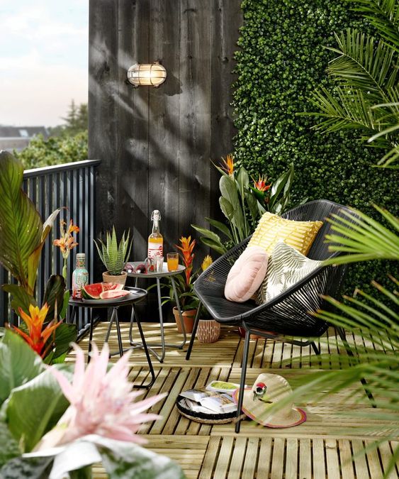 a bright balcony with a wooden floor, a living wall, round tables, a black chair and bright pillows plus potted blooms and greenery