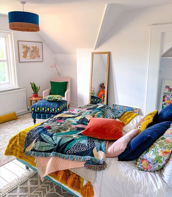 a bright bedroom with a pink chair, a colorful ottoman, bold bedidng with various prints and a green fringe pendant lamp