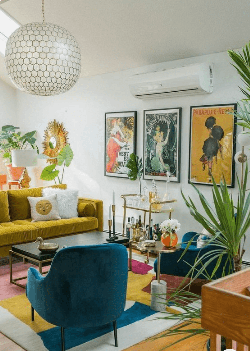 a bright living room with a mustard sofa, blue chairs, a colorful rug and a bold gallery wall plus potted plants
