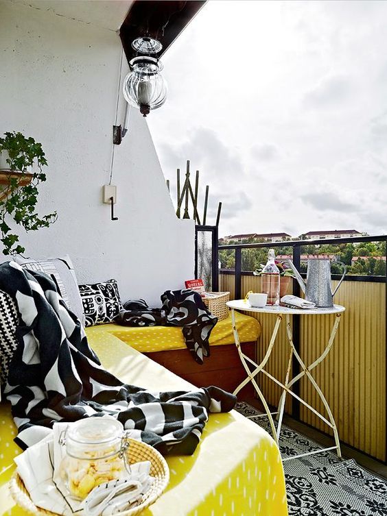 a bright summer balcony with a corner bench and yellow, black and white textiles, a shabby chic table and a potted plant 