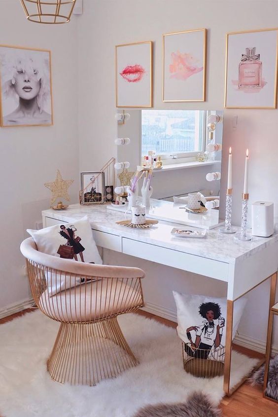 a lovely glam makeup space with a white vanity, a brass and pink chair, a glam gallery wall and a lit up mirror
