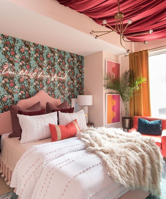 a pretty bedroom with a floral wallpaper wall, a neon sign, a pink canopy on the ceiling, a coral chair and bright artworks