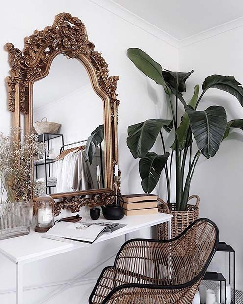 a refined makeup spot with a white vanity, a rattan chair, a mirror in a heave vintage frame, a candle and a potted plant