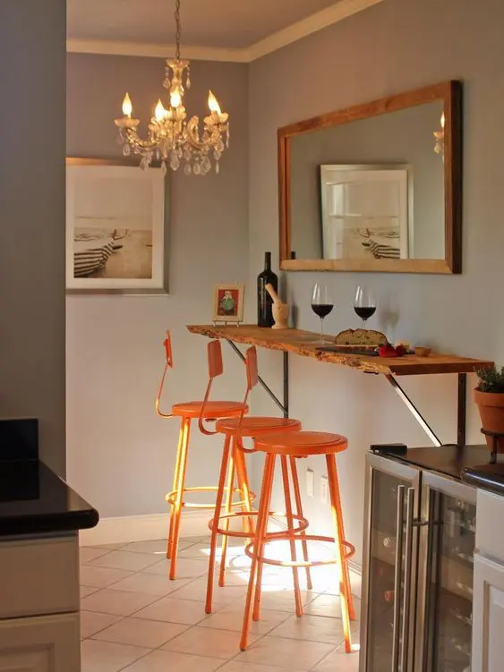 a small and chic breakfast bar with a mirror, a living edge breakfast bar, orange stools, a chic chandelier
