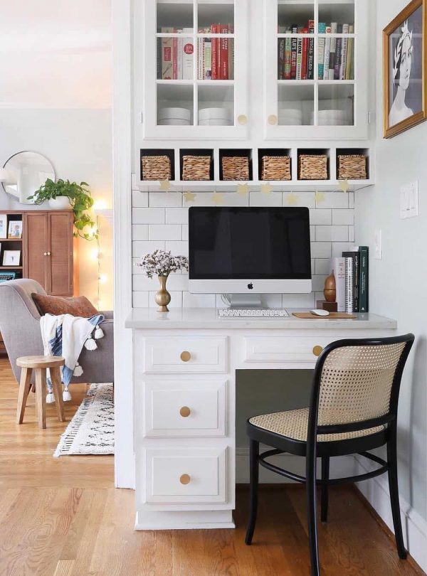 a white workspace in the kitchen with a tiny desk and a rattan chair, a glass cabinet and boxes that match the kitchen style