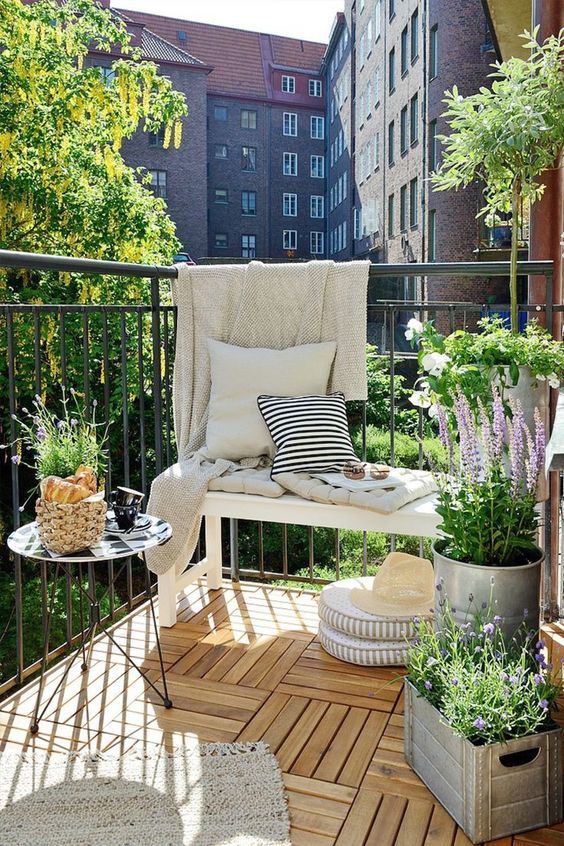an airy summer balcony with a white bench and printed textiles, a small table, potted blooms and greenery and cushions