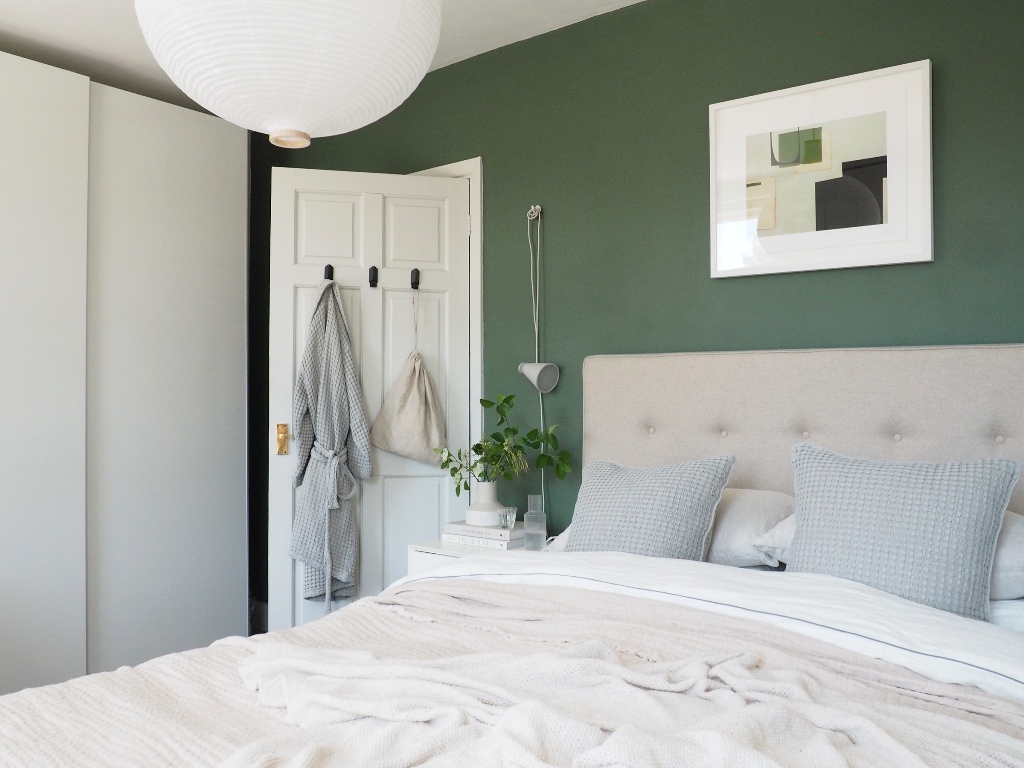 a bedroom with green walls and neutral furniture, a green artwork and potted greenery plus a paper lamp