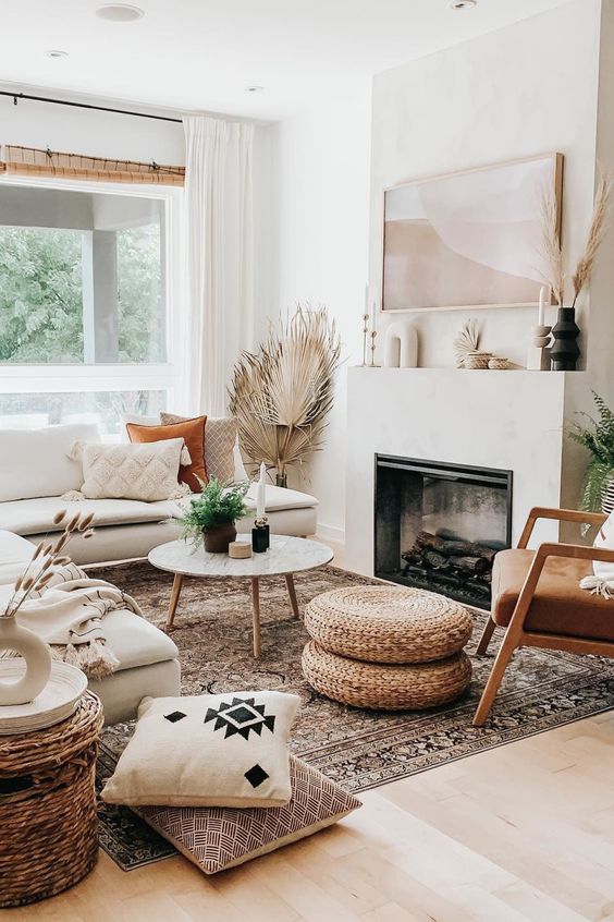 a boho summer living room with a built-in fireplace, a creamy sectional, a leather chair, woven poufs, printed pillows