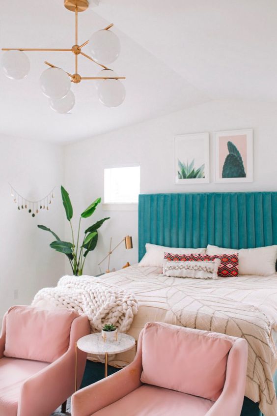 a cheerful modern bedroom with a blue upholstered bed, pink chairs, neutral textiles, a chandelier and a potted plant