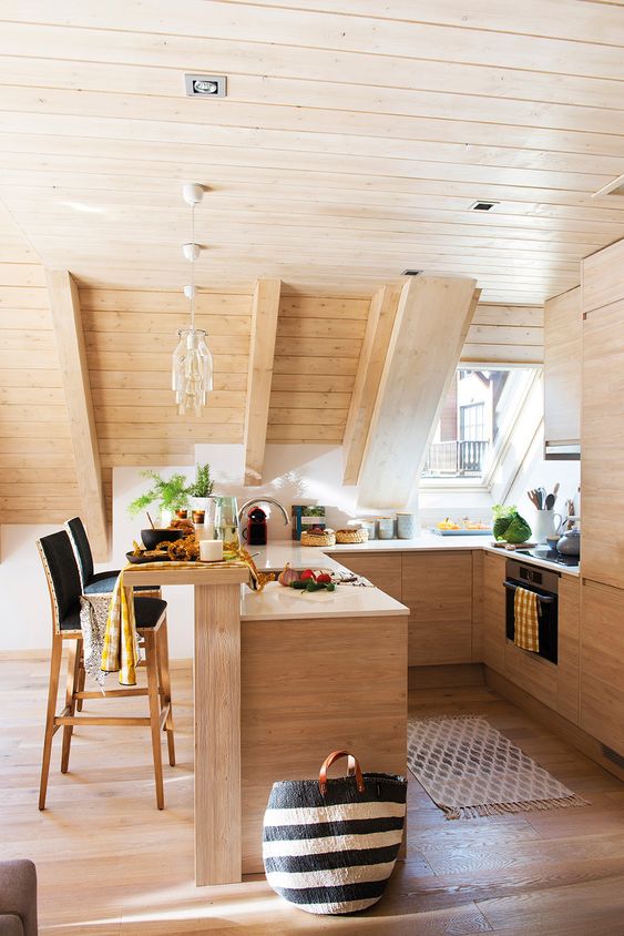 a cozy attic kitchen clad with wood, with light stained cabinetry, pendant lamps, black stools and skylights