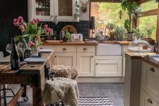 a cozy small kitchen with large windows and a skylight, vintage ivory cabinetry, black square tiles, a rough wooden table and lots of plants
