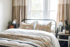 a cozy summer bedroom with a metal bed and mismatching nightstands, neutral printed textiles and potted greenery