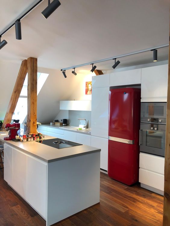a minimalist attic kitchen with sleek white cabinetry, a bold red fridge, wooden beams and lights over the spac