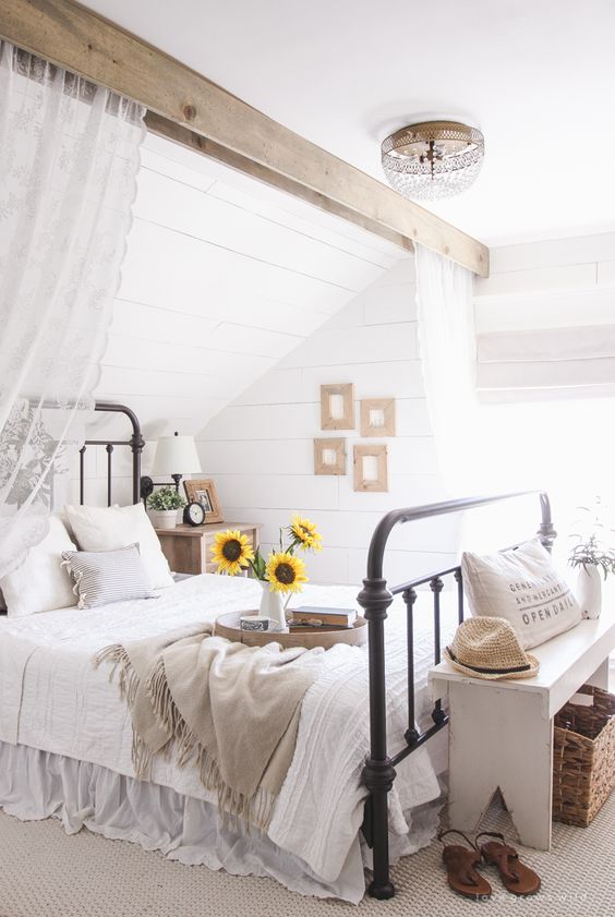 a rustic summer bedroom with a wooden beam with curtains, a metal bed with neutral bedding, a bench with decor and a small gallery wall