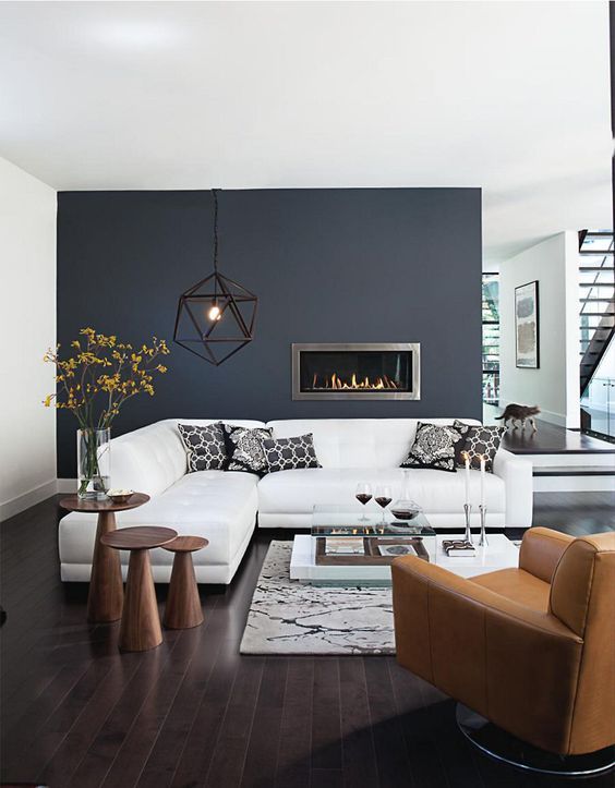 a stylish modern living room with a navy accent wall and a built-in fireplace, a white sectional, an amber leather chair and wooden round tables