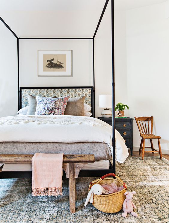 a summer bedroom with a canopy bed with bright pillows and upholstery, a dark nightstand, a wooden bench and chair and a printed rug