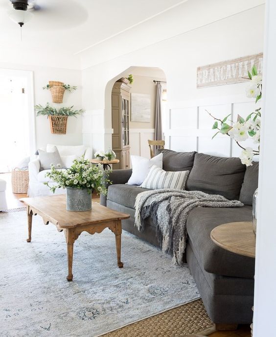 a summer cottage living room with a graphite grey sofa, white chairs, printed pillows, potted plants, layered rugs and a wooden coffee table