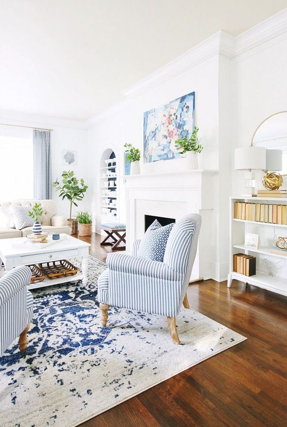 a summer living room with a fireplace, a creamy sofa, a low table, striped chairs, some potted plants and a watercolor artwork