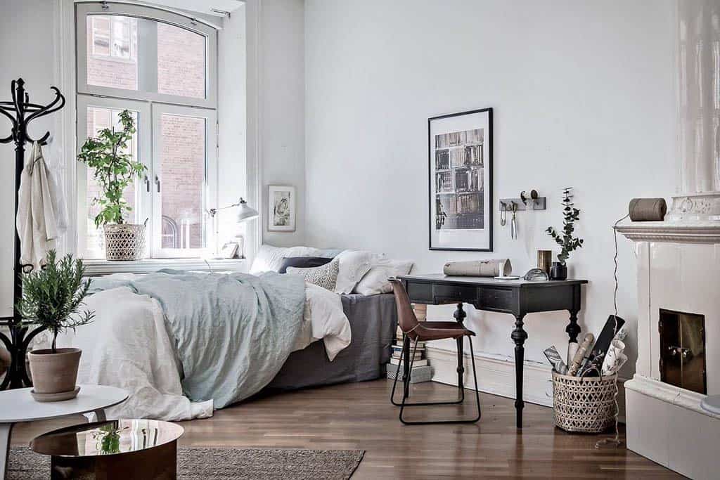a vintage Scandinavian bedroom with a stove, a bed with pastel bedding, a dark desk with a leather chair and potted plants