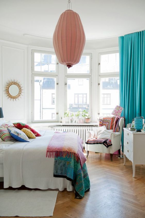 a vivacious summer bedroom with a bow window, neutral furniture, colorful printed textiles, a pink pendant lamp and turquoise curtains