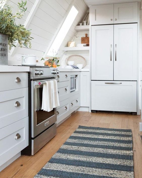 a white farmhouse attic kitchen with shaker style cabinets and planked walls, potted plants and a striped rug