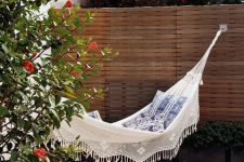 16 a chic outdoor space with greenery and blooms, a white hammock with printed pillows, a small jute pouf and a glass of wine