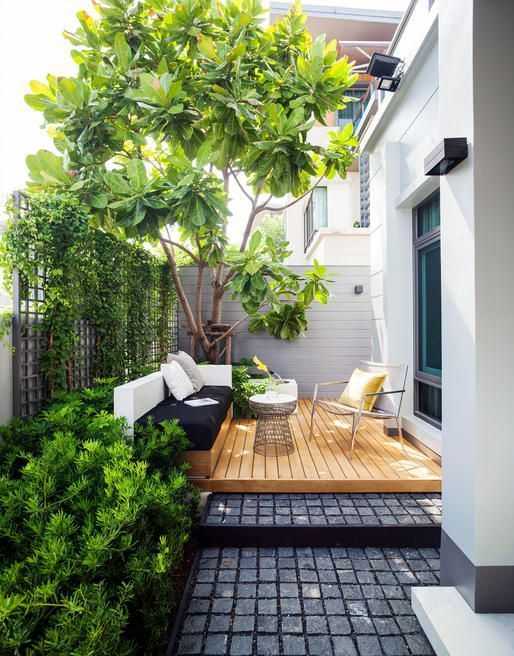 a small contemporary backyard with a wooden deck, a built-in daybed and a cool chair, some growing plants and a large living tree