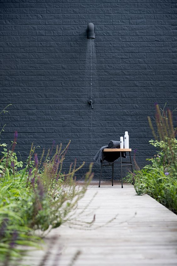 a Nordic outdoor shower with a black brick wall, a small stool with towels and shampoos, a wooden deck