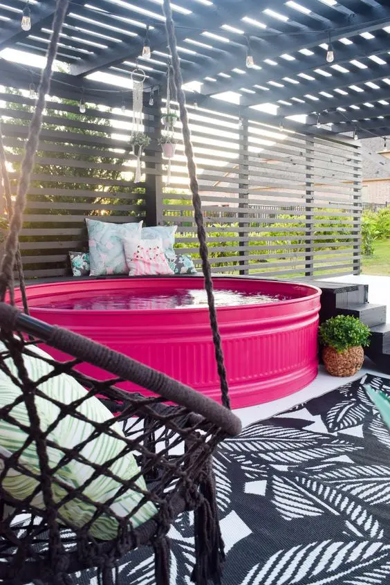 a bold patio with a hot pink stock tank pool, a black ladder, a woven chair with a pillow and some more pillows on the bench