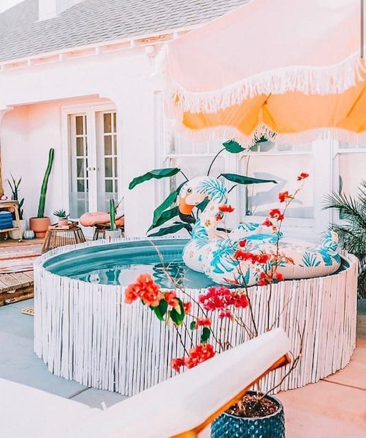 a bright and cool backyard with a wooden deck, a stock tank pool covered with whitewashed sticks, a colorful float, an umbrella and bold potted blooms