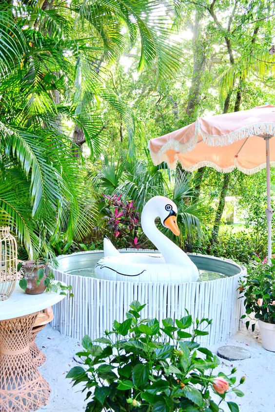 a bright and funny pool nook with a stock tank pool clad with a stick cover, potted greenery all around, a pink umbrella and rattan furniture