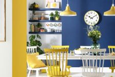 a chic and bold dining room with a navy wall, a yellow ceiling, chairs, pendant lamps and pillows plus navy built-in shelves