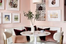 a chic dining nook with blush walls, a built-in white bench, a white round table and elegant white and brass chairs plus a cool gallery wall