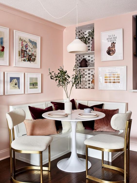 a chic dining nook with blush walls, a built-in white bench, a white round table and elegant white and brass chairs plus a cool gallery wall