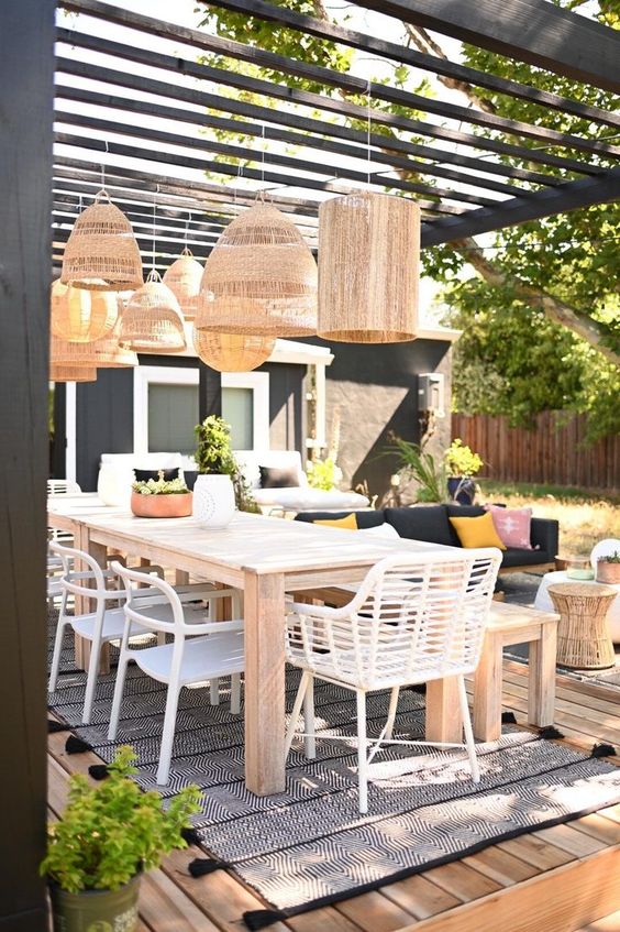 a cool modern outdoor dining space with a stained table and a bench, white chairs, woven pendant lamps and a sitting zone next to it