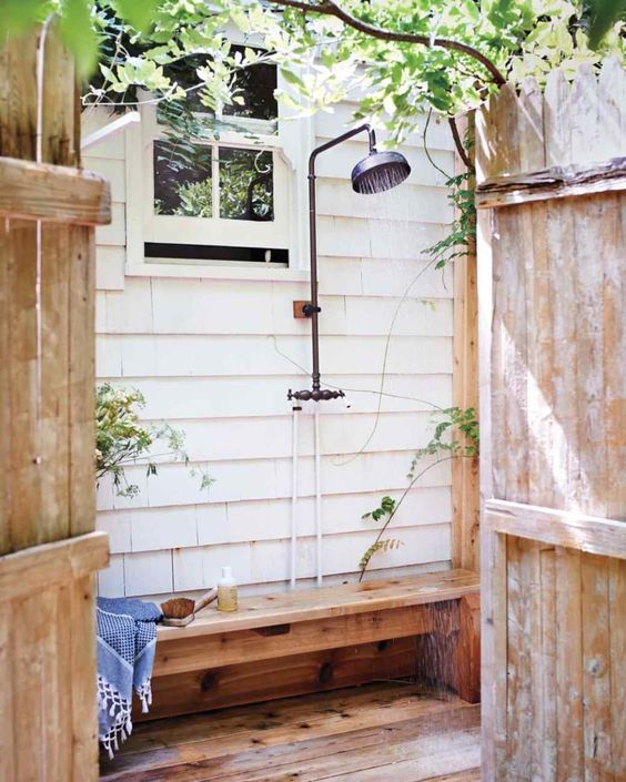a cozy farmhouse shower space with a planked wall, a stained wooden floor and a bench, greenery around and some necessary stuff