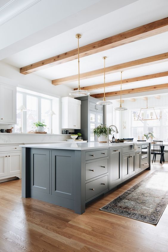a farmhouse kitchen with white cabinets and a slate grey ktichen island, wooden beams and lovely plate pendant lamps plus wooden beams