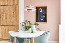 a lovely dining space with a blush accent wall, a white table and blue chairs, a pink pendant lamp and a statement potted plant