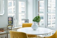 a lovely modern dining space with a view, a round hairpin table, yellow chairs, a gilded bar cart, a chic vase with a leaf