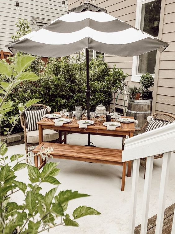 a lovely outdoor dining nook with a simple planked wood dining set, rattan chairs, a striped umbrella and lots of greenery around