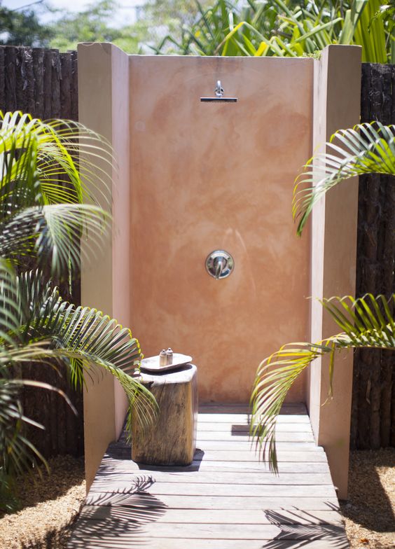 a lovely outdoor shower with a wooden deck, pink walls and a tree stump stool plus plants around is a cool space to refresh yourself