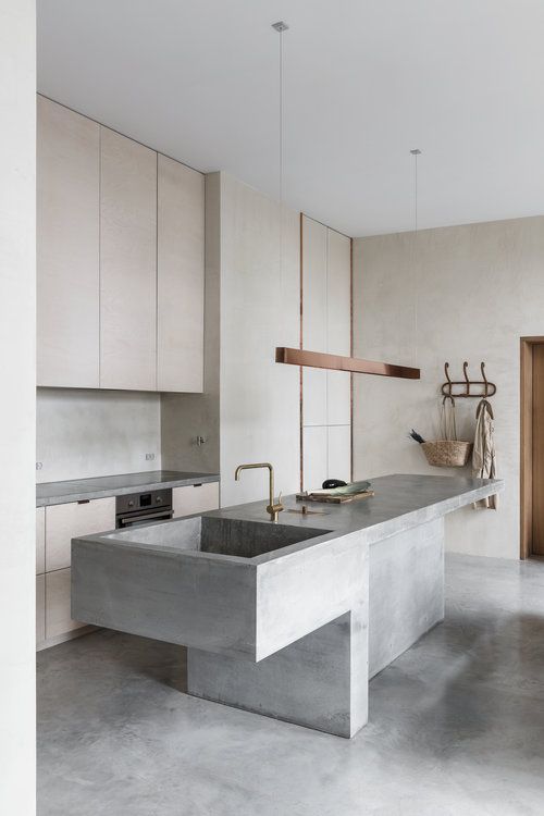 a minimalist kitchen with light stained cabinets, a concrete backsplash and a kitchen island with a sink, a chic pendant lamp