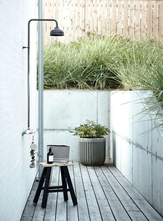 a modern outdoor shower with a wooden deck, a stool, potted plants, a shampoo and lots of grass growing around