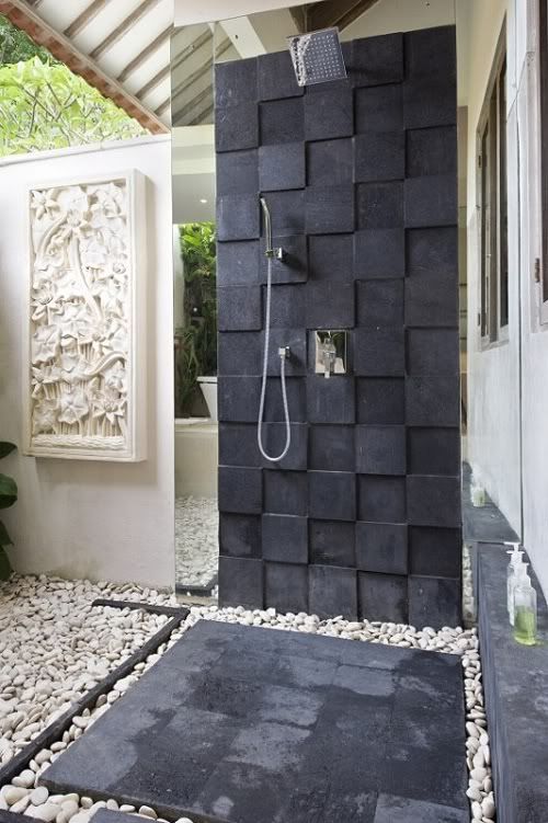a modern outdoor shower with pebbles on the ground, with black tiles on the wall and ground, plus a roof over it