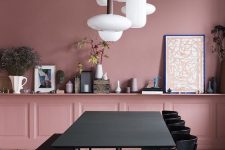 a modern pink dining room with pink paneled walls, a long shelf along the whole wall, a black dining set and a cluster of pendant lamps