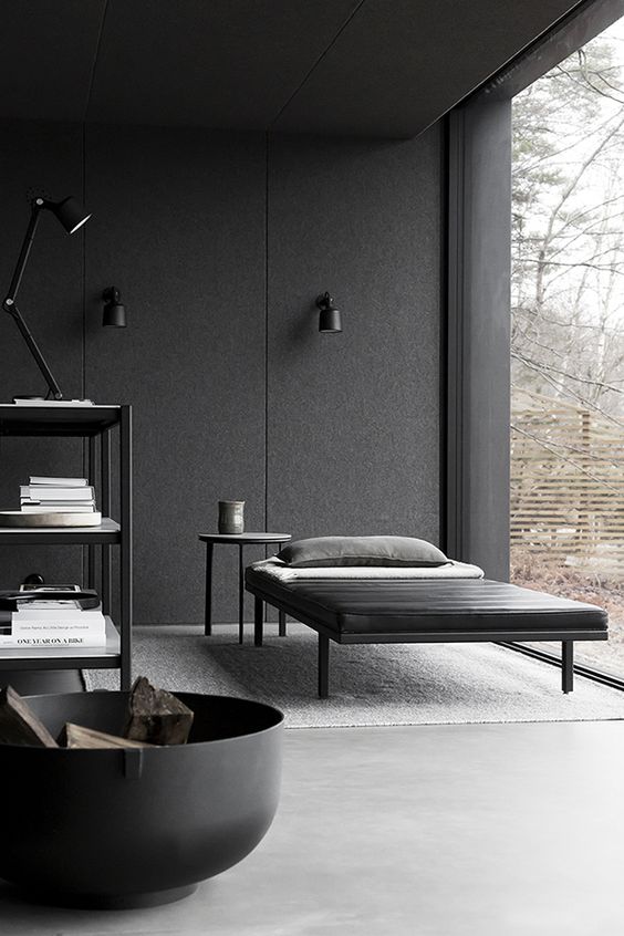 a moody minimalist living room with a glazed wall, a black daybed and side table, a fire pit and a shelving unit plus lamps