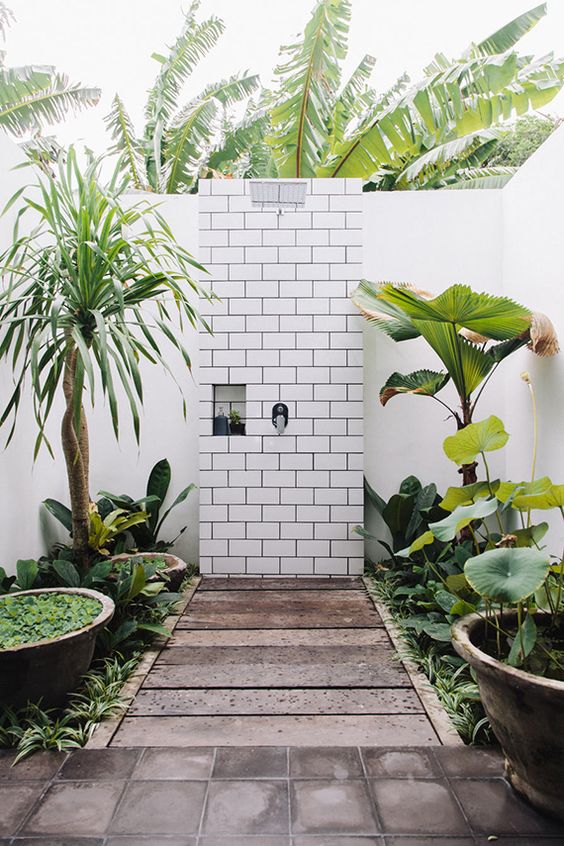 a pretty outdoor bathroom with a wooden deck and a subway tile wall, growing plants around is a very lovely space