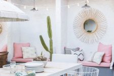 a pretty pastel summer terrace with a built-in grey bench and pink upholstery, a white table and white chairs, a potted cactus and a ligth string