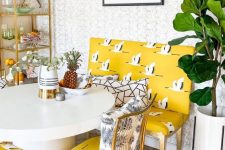 a quirky dining room with a gilded storage unit, a round table, whimsical yellow printed chairs and a loveseat plus a potted plant